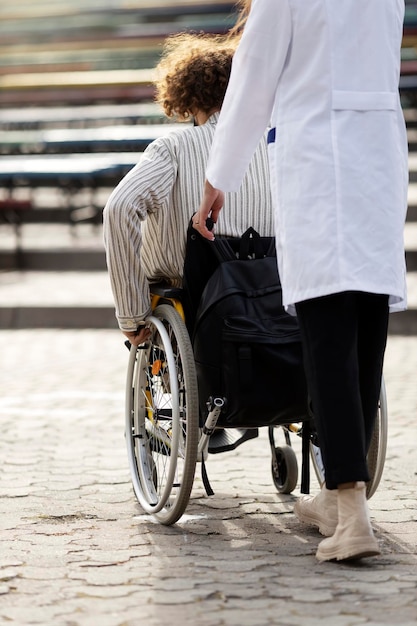 Enfermera de vista trasera que lleva una silla de ruedas con un paciente Asistencia a un paciente con discapacidad al aire libre