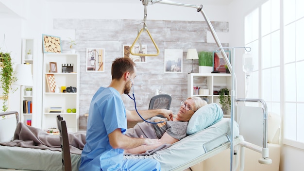 Enfermero comprobando los latidos del corazón de una anciana enferma acostada en la cama de un hospital en un hogar de ancianos luminoso y acogedor