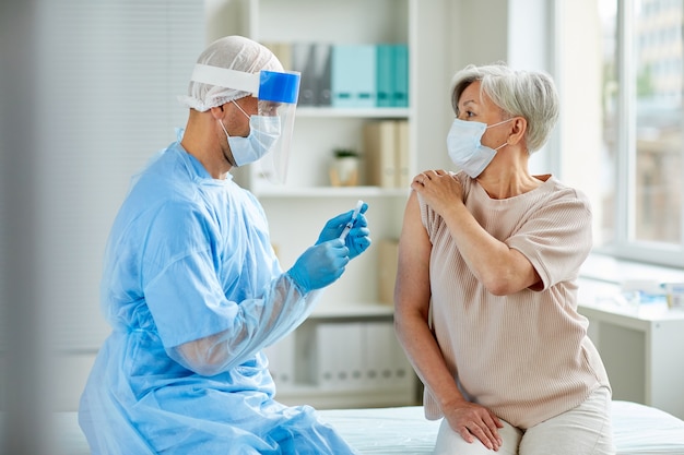 Enfermero con máscara protectora y guantes preparando una jeringa médica para administrar la inyección a un paciente mayor