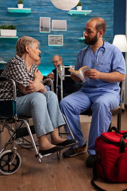 Enfermero que brinda apoyo al paciente leyendo historias de libros a ancianas con discapacidad en la sala de estar. Servicios sociales de enfermería a personas mayores en centro de salud.