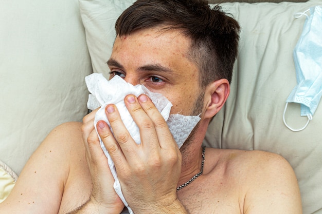 Enfermo sufre de gripe viral en la cama, se sopla la nariz con un pañuelo desechable