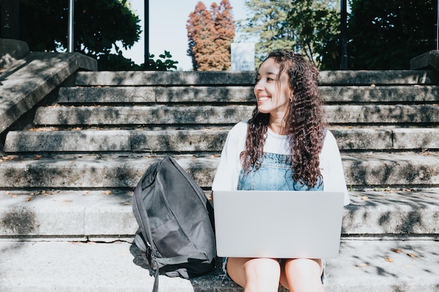 Enfocado joven estudiante afroamericano feliz sentado en las escaleras en la universidad usando una computadora portátil para investigar sobre trabajos universitarios y papel. Educación: tomar notas, conferencias en línea, adquirir conocimientos