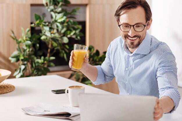 Enfoque positivo del trabajo. Autónomo autónomo sonriente creativo dotado sentado en casa y bebiendo jugo mientras usa gadget y trabaja