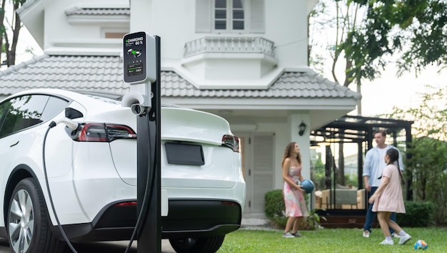 Enfoque de recarga de vehículos eléctricos desde la estación de carga doméstica en un fondo borroso de una familia feliz y juguetona jugando juntos en un coche eléctrico utilizando energía alternativa y sostenible para un futuro mejorSynchronos