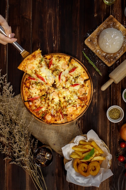 Enfoque selectivo y cierre un pedazo de pizza en una espátula o una cuchara con la mano sosteniendo o presentando con desenfoque de fondo de madera y decorado con tela de saco, ingredientes, papas fritas y bebida fría.