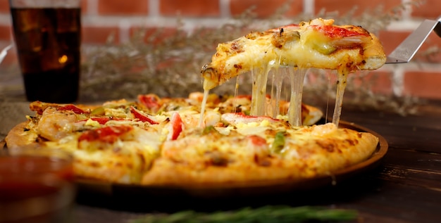 Enfoque selectivo y cierre en un trozo de pizza casera de mariscos con doble queso en una espátula o cuchara con fondo borroso de mesa de madera, bebida fría y pared de ladrillo. Concepto de comida.