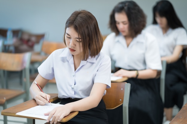 El enfoque selectivo de los estudiantes universitarios adolescentes sentados en la silla de conferencias en el aula escribe en la hoja de respuestas del papel de examen al hacer la prueba del examen final. Alumnas en uniforme de estudiante.