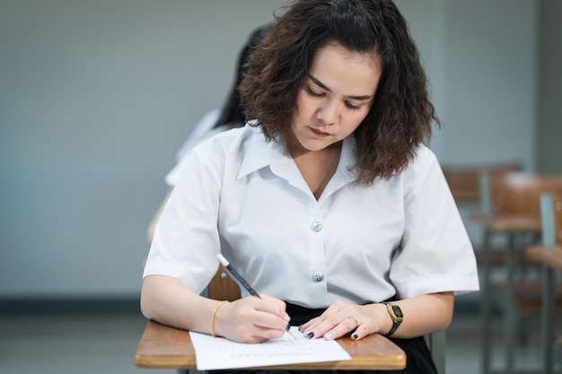 El enfoque selectivo de los estudiantes universitarios adolescentes se sientan en la cátedra de conferencias, escriben en la hoja de respuestas del papel de examen y toman la sala de examen final o el aula. Estudiantes universitarios en uniforme en el aula.