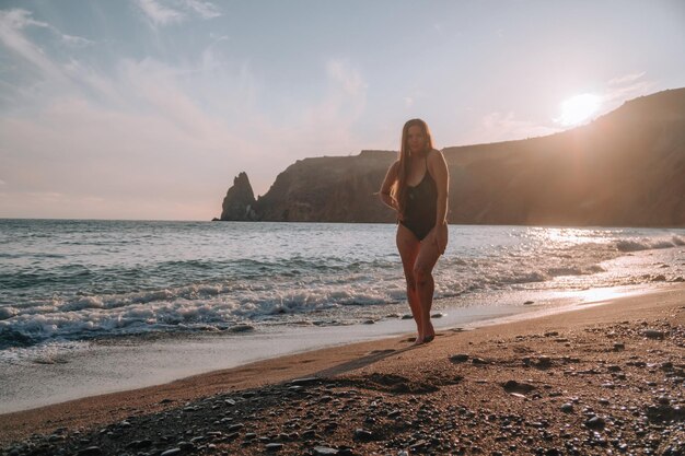 Enfoque selectivo feliz mujer sensual despreocupada con cabello largo en traje de baño negro posando en la playa al atardecer