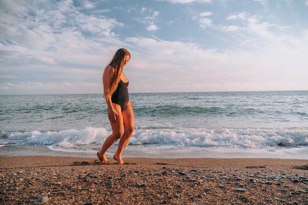 Enfoque selectivo feliz mujer sensual despreocupada con cabello largo en traje de baño negro posando en la playa al atardecer