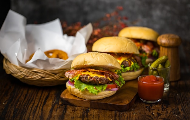 Enfoque selectivo en hamburguesas con hamburguesas de ternera, cebollas fritas, espinacas, salsa de tomate, pimiento y queso acompañadas de guarniciones en una tabla de madera con refresco.