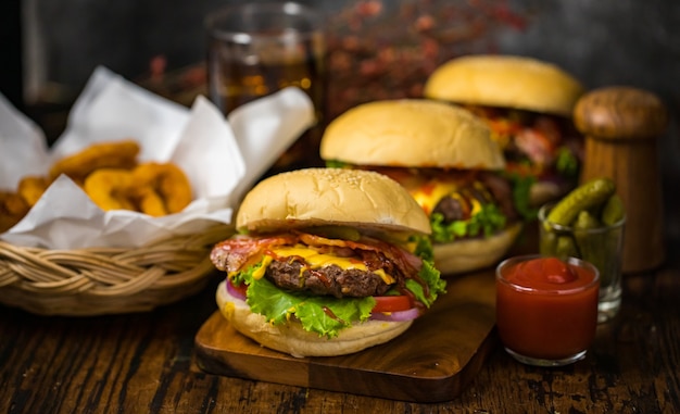 Enfoque selectivo en hamburguesas con hamburguesas de ternera, cebollas fritas, espinacas, salsa de tomate, pimiento y queso acompañadas de guarniciones en una tabla de madera con refresco.