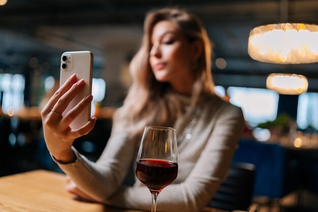 Enfoque selectivo de una joven borrosa sentada en la mesa con una copa de vino tinto usando un teléfono móvil en vivo en las redes sociales escribiendo historias tomando selfies