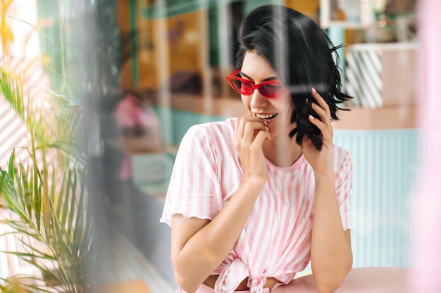 Enfoque selectivo de la juguetona chica europea en gafas de sol rosas Toma al aire libre de la alegre dama morena en camiseta rosa