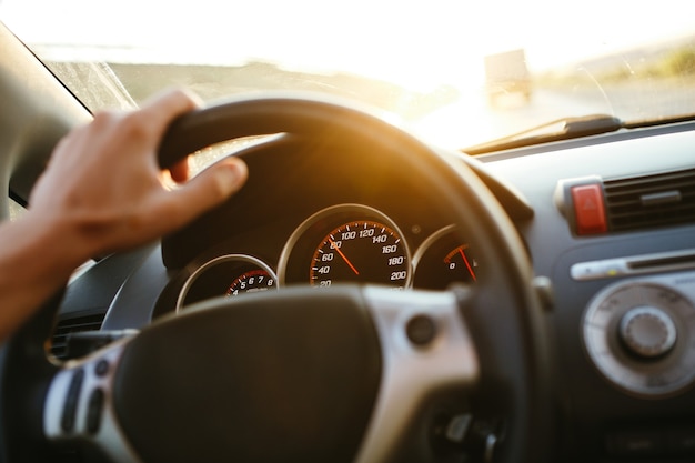 Enfoque selectivo de la mano del hombre en el volante, conduciendo un coche al atardecer. Antecedentes de viaje.