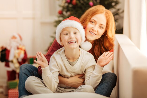 Enfoque selectivo en un niño extremadamente feliz que se emociona mientras está sentado junto a su amorosa madre abrazándolo en un sofá.