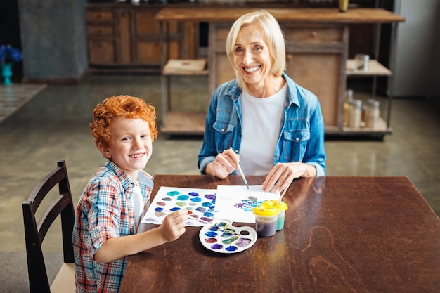 Enfoque selectivo en un pequeño artista de pelo rizado sentado junto a su abuela mientras pintan y miran a la cámara con una sonrisa alegre en sus rostros.
