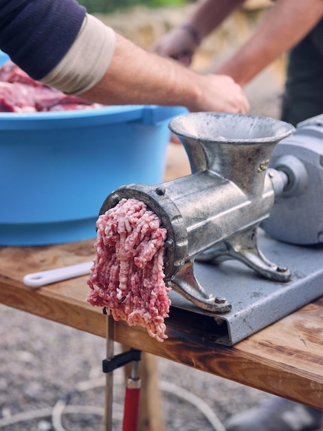 Enfoque selectivo de picadora de metal con carne picada sobre mesa de madera contra machos de cultivo con tazón de carne picada en la naturaleza