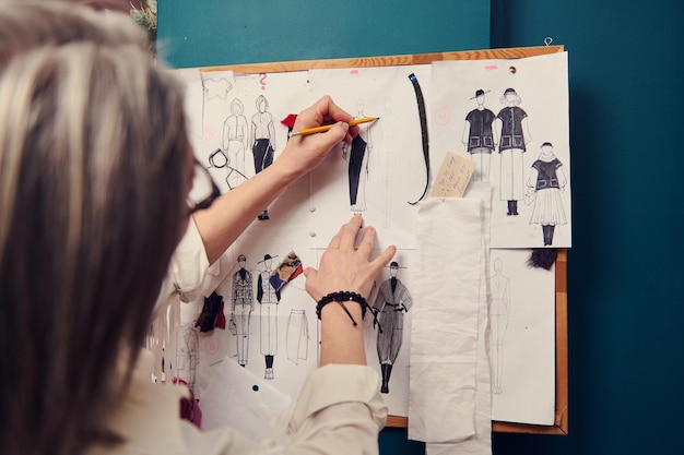 Enfoque suave en las manos del diseñador de moda sosteniendo un lápiz y esbozando ropa en papel clavado en la pared con dibujos y esquises adjuntos Concepto creativo de ocupación profesional