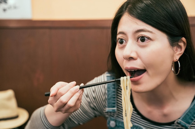 Enfóquese en la cara de una joven y hermosa chica asiática que viaja a osaka, Japón, y prueba la comida japonesa a la hora del almuerzo. mujer turista comiendo fideos con palillos. boca abierta femenina con sabroso udon en el restaurante.