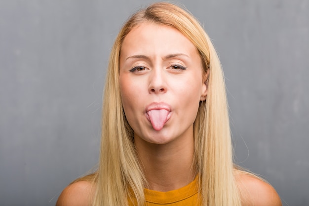 Enfrentar closeup, retrato de uma expressão natural jovem mulher loira de confiança e emoção, diversão e amigável, mostrando a língua como um sinal de jogo ou diversão