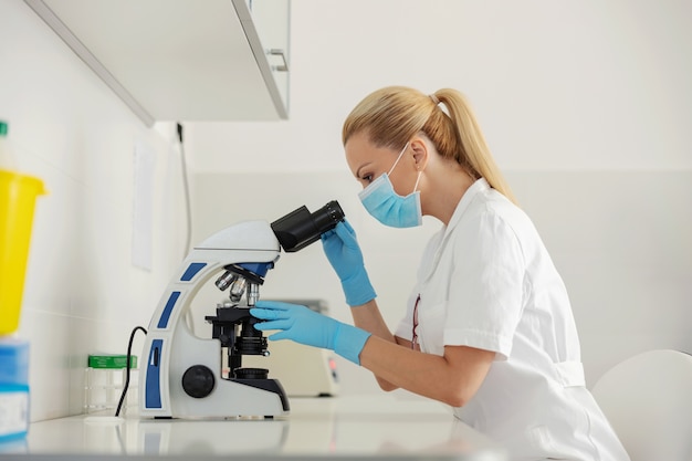 Engagierte blonde Laborassistentin, die im Labor sitzt und ein Mikroskop für die Blutuntersuchung verwendet.
