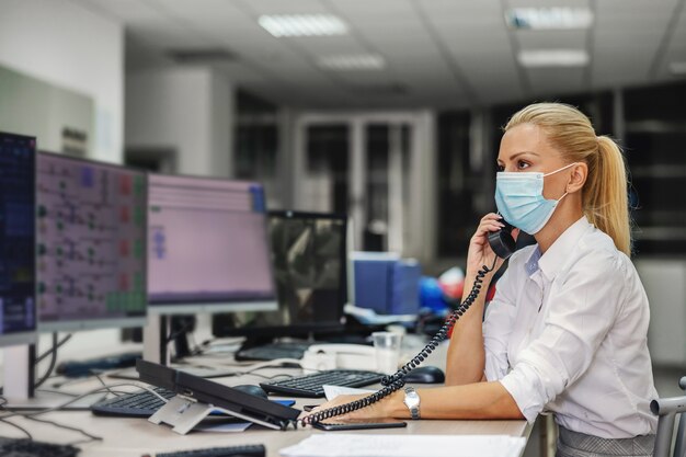 Engagierte fleißige blonde Chefin im Anzug mit Gesichtsmaske beim Sitzen im Kontrollraum im Heizwerk und bei wichtigen Telefongesprächen während des Ausbruchs des Koronavirus.