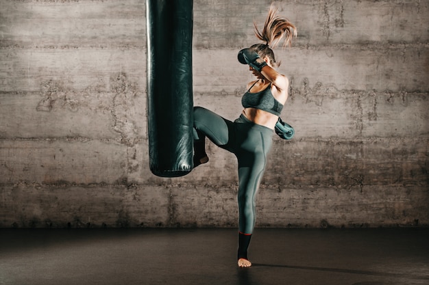 Engagierte starke Brünette mit Pferdeschwanz, in Sportbekleidung, nacktem Fuß und mit Boxhandschuhen, die im Fitnessstudio gegen den Sack treten.