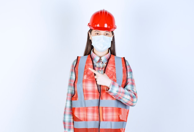 Foto engenheira civil feminina com capacete vermelho e máscara de segurança apontando para a esquerda.