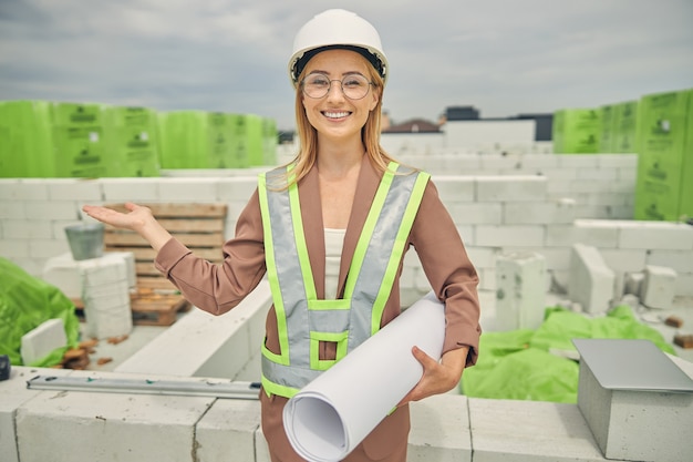 Engenheira civil loira sorridente com um capacete de segurança convidando clientes para um novo objeto de construção