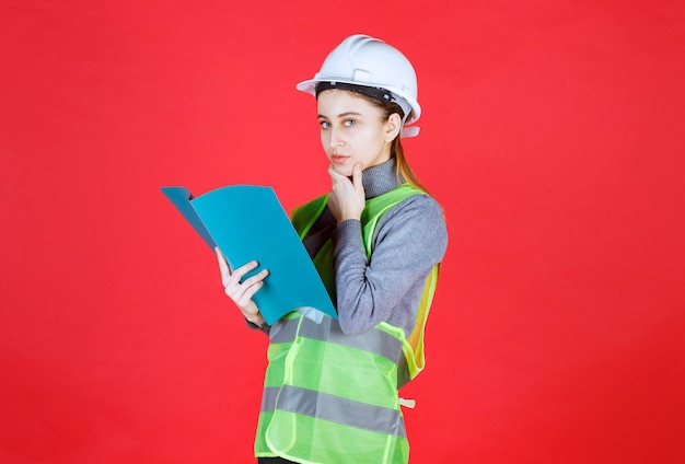 Engenheira com capacete branco, segurando um arquivo de projeto azul, lendo e pensando.