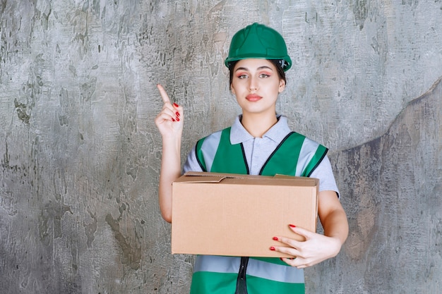 Engenheira com capacete verde segurando uma caixa de papelão e apontando para alguém ao redor