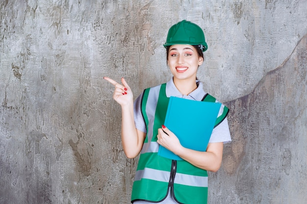 Engenheira com capacete verde segurando uma pasta azul e apontando para alguém ao redor