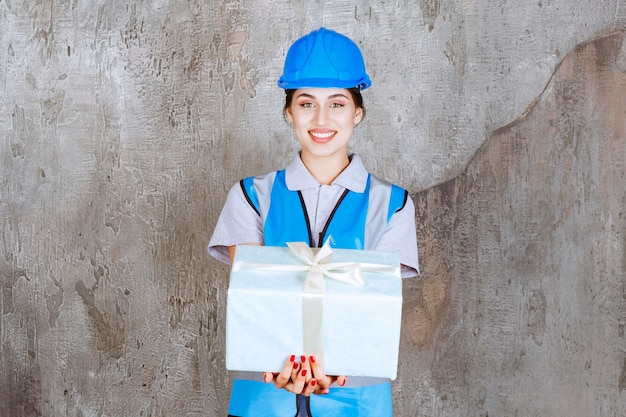 Engenheira de uniforme azul e capacete segurando uma caixa de presente azul.