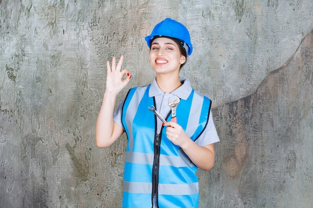 Engenheira de uniforme azul e capacete segurando uma chave metálica e mostrando sinal positivo com a mão