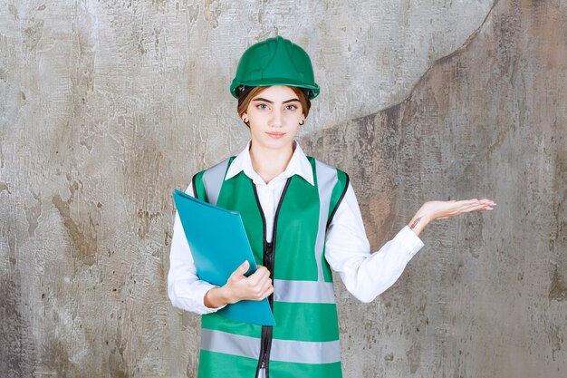 Engenheira de uniforme verde e capacete segurando uma pasta de projeto verde e apontando para o lado direito