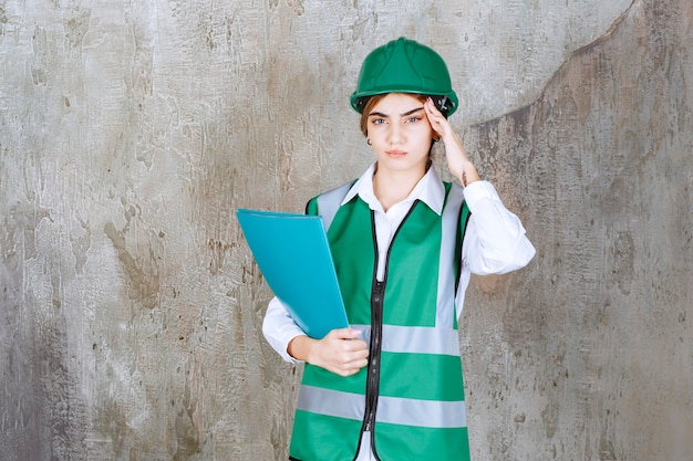 Engenheira de uniforme verde e capacete segurando uma pasta de projeto verde e parece cansada e com sono