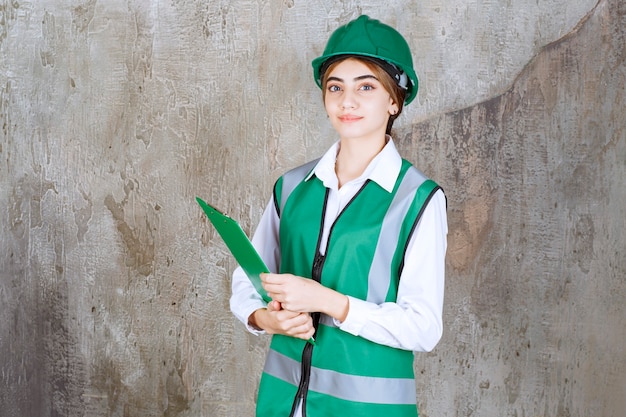 Engenheira de uniforme verde e capacete segurando uma pasta de projeto verde