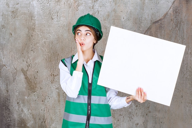 Engenheira de uniforme verde e capacete segurando uma placa retângulo de informações e parece surpresa ou animada.