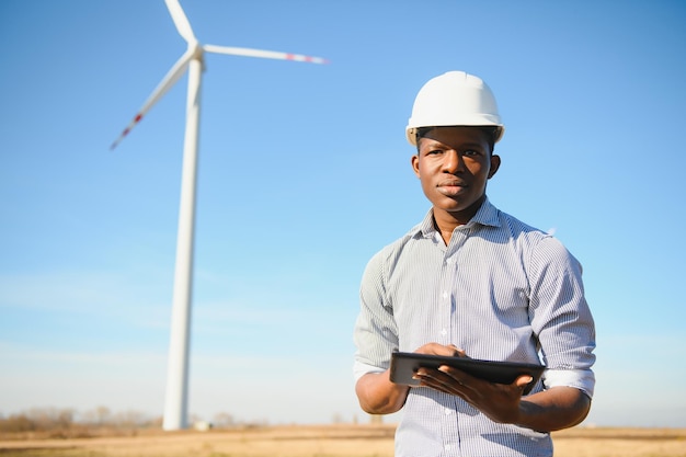 Engenheiro africano usando capacete branco em pé com tablet digital contra turbina eólica