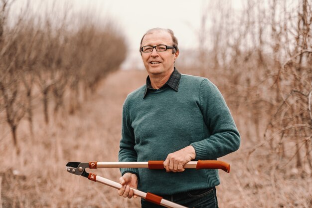 Engenheiro agrônomo sênior caucasiano com óculos, segurando uma tesoura de poda e em pé no pomar.