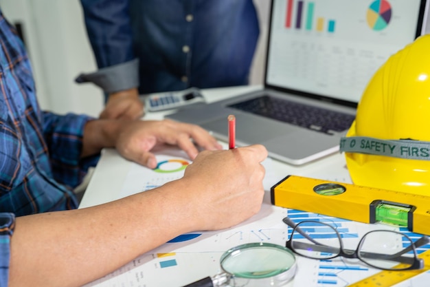 Engenheiro asiático e equipe de parceiros aprendem e conversam com o gráfico de planta do projeto de plano de casa e capacete amarelo e ferramentas de construção de engenharia no trabalho de mesa