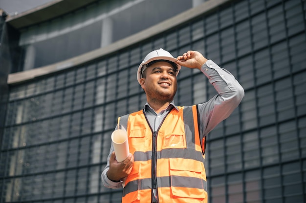 Engenheiro asiático homem bonito ou arquiteto ansioso segurando a planta de papelada com capacete de segurança branco no canteiro de obras em pé no edifício moderno