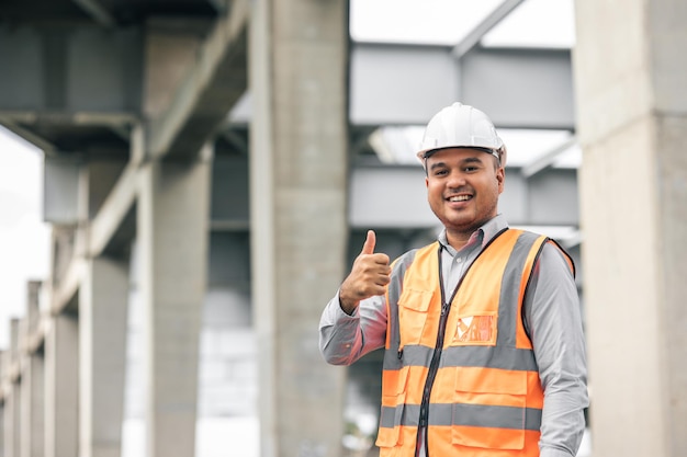 Foto engenheiro asiático homem bonito ou arquiteto procurando construção com capacete de segurança branco no canteiro de obras em pé no local da estrada de concreto da rodovia mostrando os polegares para cima