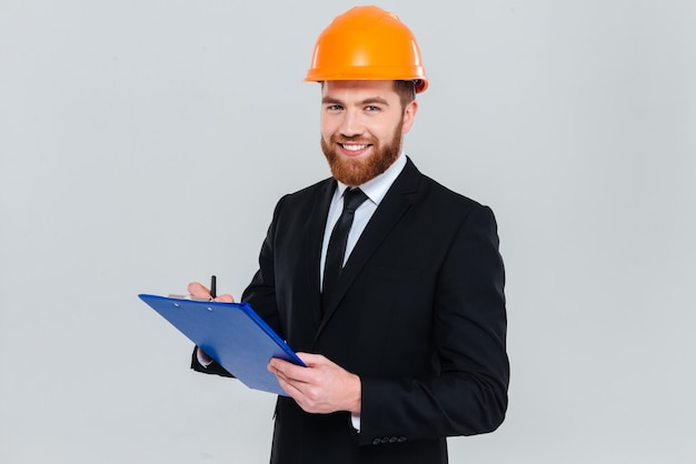 Engenheiro barbudo sorridente de terno e capacete segurando uma prancheta