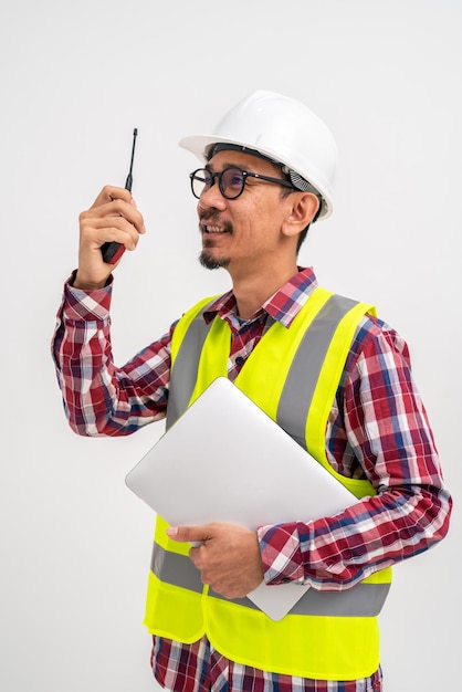 Engenheiro civil barbudo asiático em capacete usando colete está usando walkie-talkie com laptop em pé sobre fundo branco Conceito de engenheiro civil de construção Imagem vertical