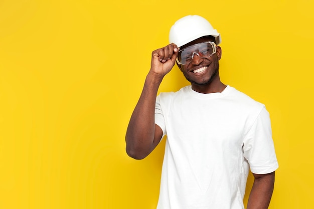 engenheiro civil masculino afro-americano com capacete e óculos sorri em fundo amarelo isolado