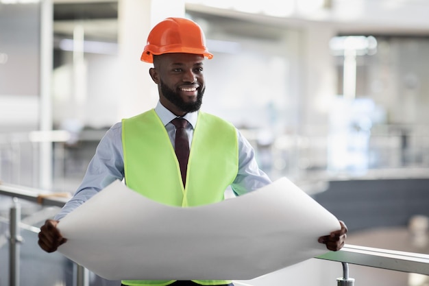 Engenheiro civil preto alegre em roupas de trabalho de segurança segurando rascunho