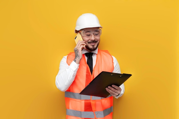 Engenheiro construtor asiático de uniforme falando ao telefone sobre fundo amarelo isolado