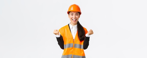 Engenheiro de gerente de construção feminino asiático vencedor bem-sucedido na construção de vista usando capacete de segurança, levantando as mãos na bomba de punho alegre da conquista e vitória no fundo branco
